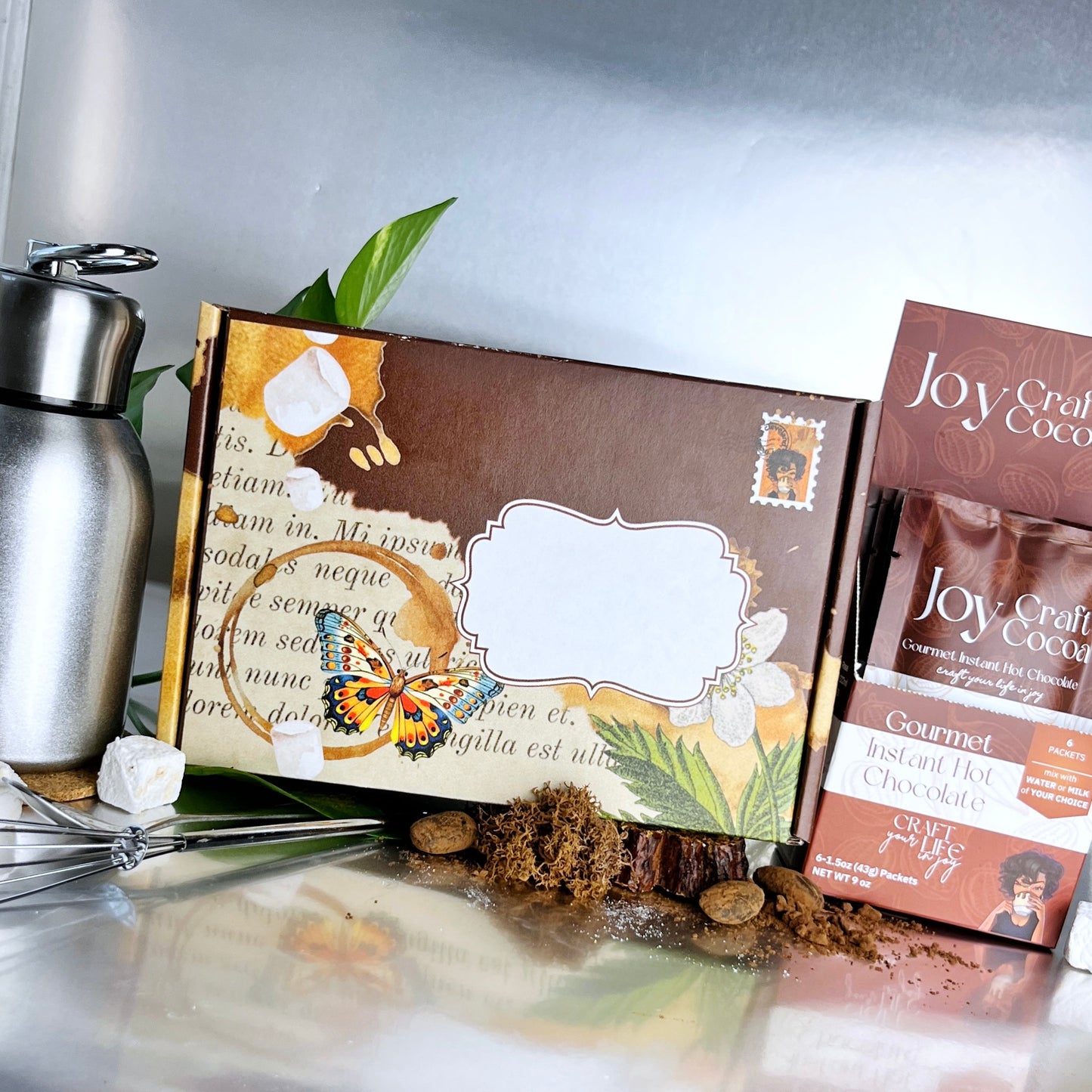 Hot Chocolate Experience Gift Box a thermos, a whisk, a spoon, a brown shipping box with a butterfly on it. a box of Joy Craft Cocoa single packs