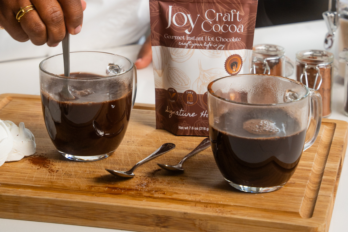 cutting board on a table with 2 cups of hot chocolate.  in the background is a bag of hot chocolate with the words joy craft cocoa